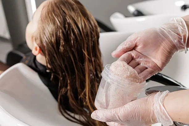 Gloved stylist prepares scalp scrub with exfoliating microgranules at a salon basin for detox, dandruff care, and hygienic hair treatment.