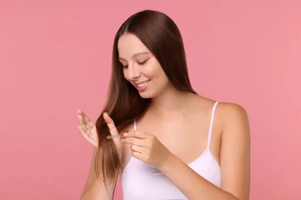 Beautiful woman applying serum onto hair on pink background