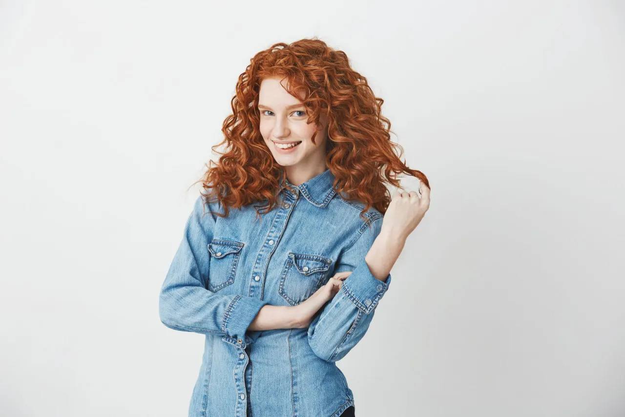 Playful beautiful redhead girl smiling touching hair looking at camera over white background. Copy space.